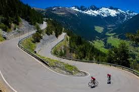 Two road cyclists on a hairpin bend of a Pyrenean mountain road, with snow-capped peaks behind them